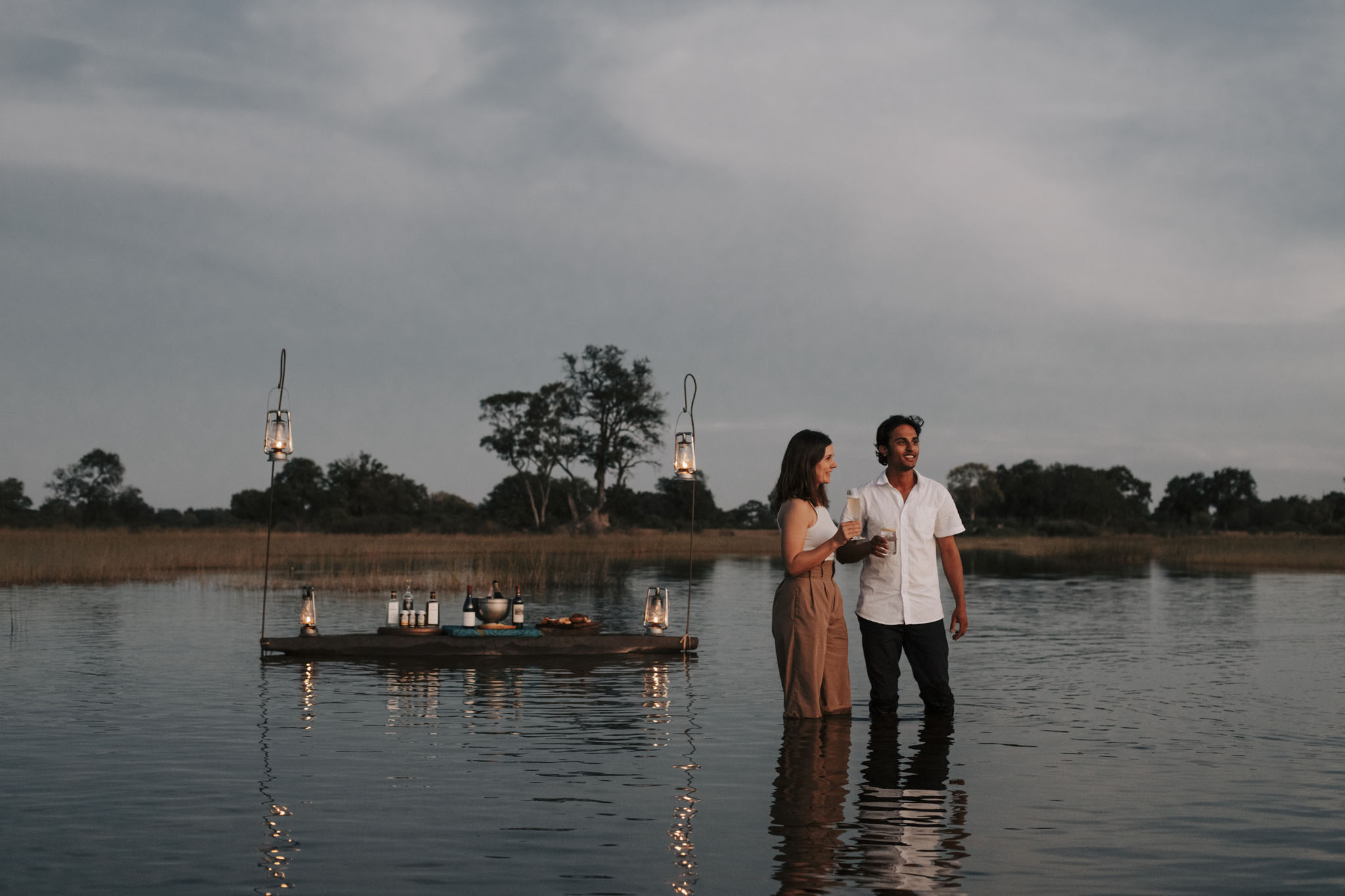 romantic dinner set up in water of delta with couple standing in water nearby at Vumbura Plains Camp Wilderness Okavango Delta